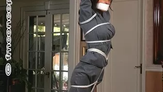 Trussed-up in her business suit and tape-gagged, Gia Mancini stands with her arms stretched overhead by a rope-link!